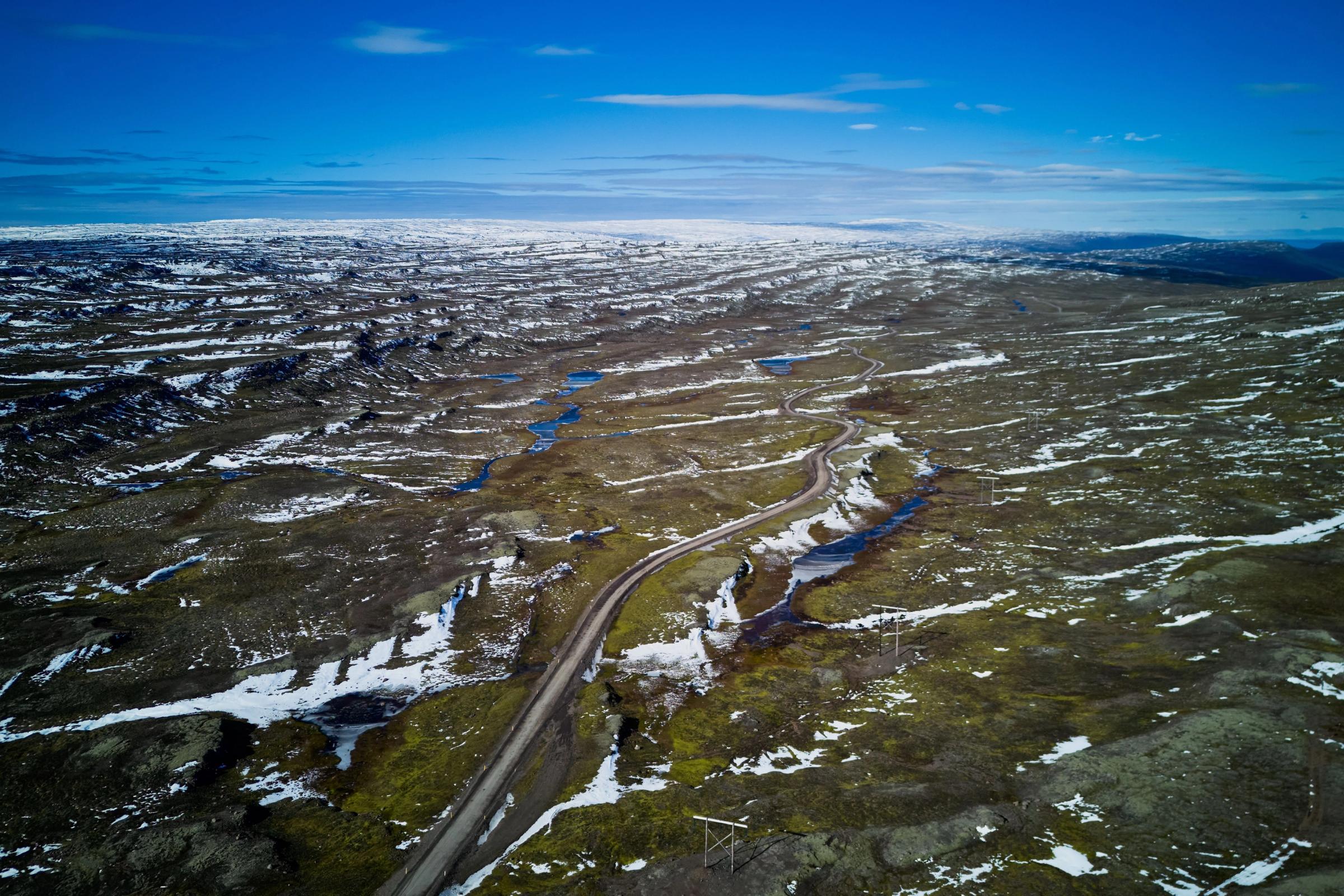 Vista aérea de una carretera serpenteante a través de un vasto paisaje parcialmente cubierto de nieve y musgo con pequeños lagos azules bajo un cielo azul.