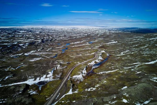 Aerial view of a winding road through a vast, partially snow-covered, mossy landscape with small blue lakes under a blue sky.