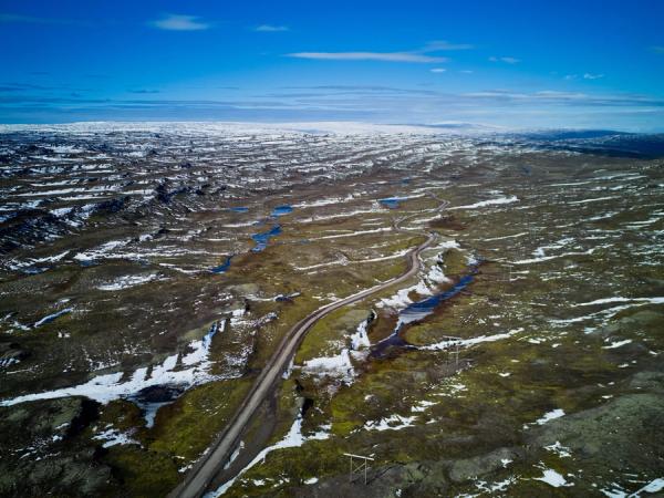 Aerial view of a vast, partially snow-covered landscape with a winding road, blue ponds, and green-brown terrain under a blue sky.