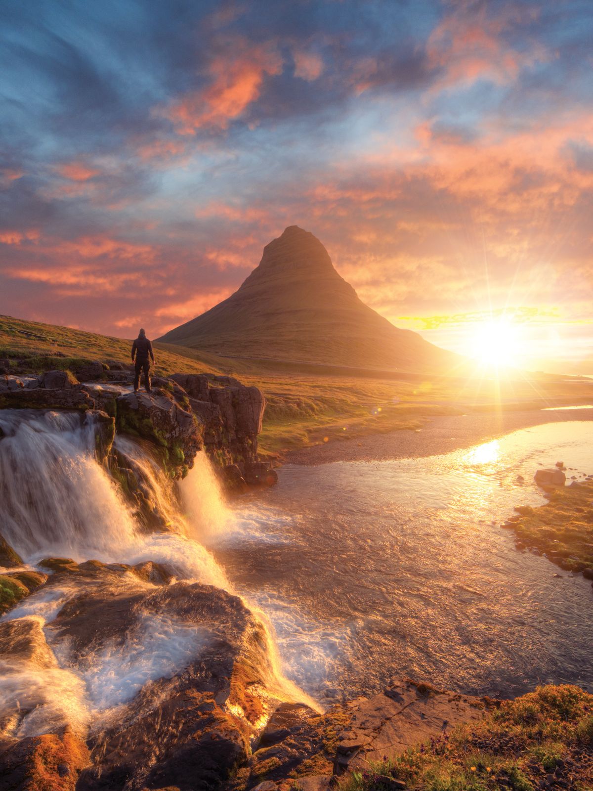 a man is standing on a rock overlooking a waterfall and a mountain at sunset .