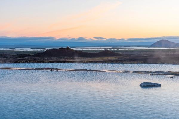 Steaming light blue geothermal water with dark volcanic hills under a pastel sunset sky.