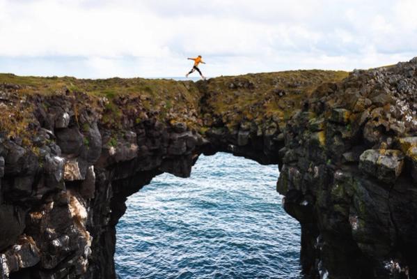 A person wearing an orange jacket jumps through a natural rock arch over the ocean.