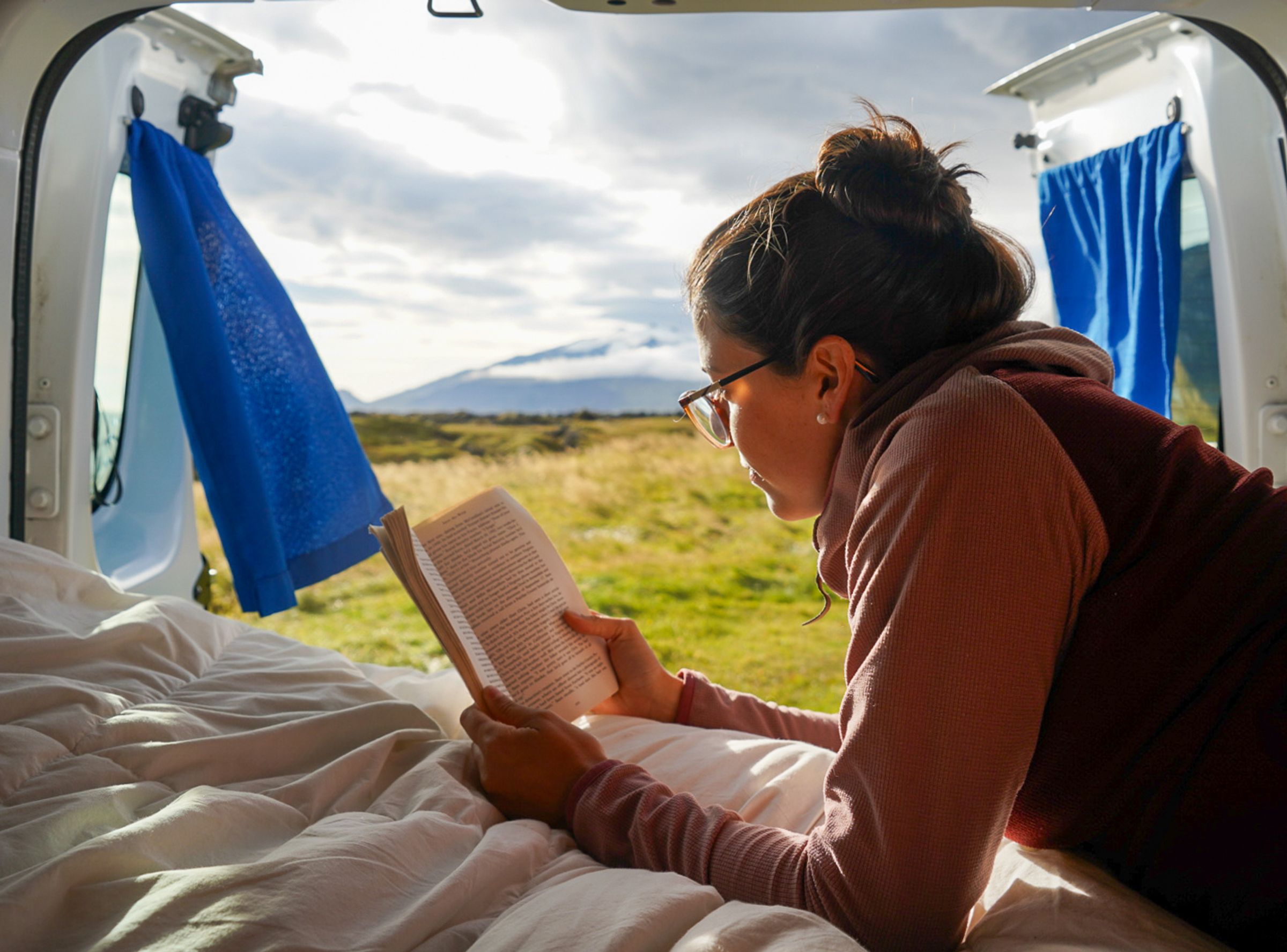 Eine Person liegt auf dem Bett und liest ein Buch, die Hecktüren sind geöffnet und geben den Blick auf eine Wiese und einen Berg frei