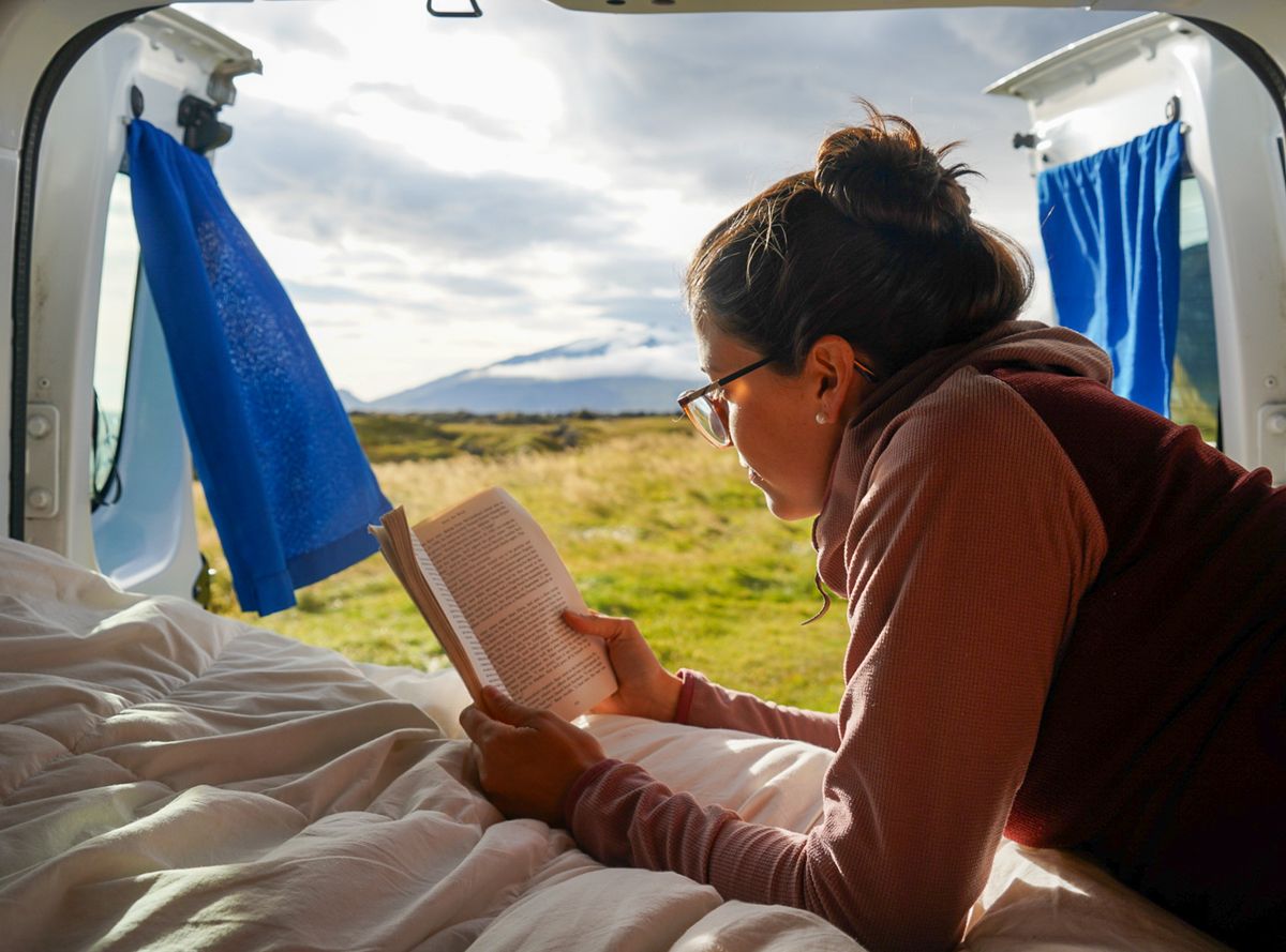 Eine Person liegt auf dem Bett und liest ein Buch, die Hecktüren sind geöffnet und geben den Blick auf eine Wiese und einen Berg frei