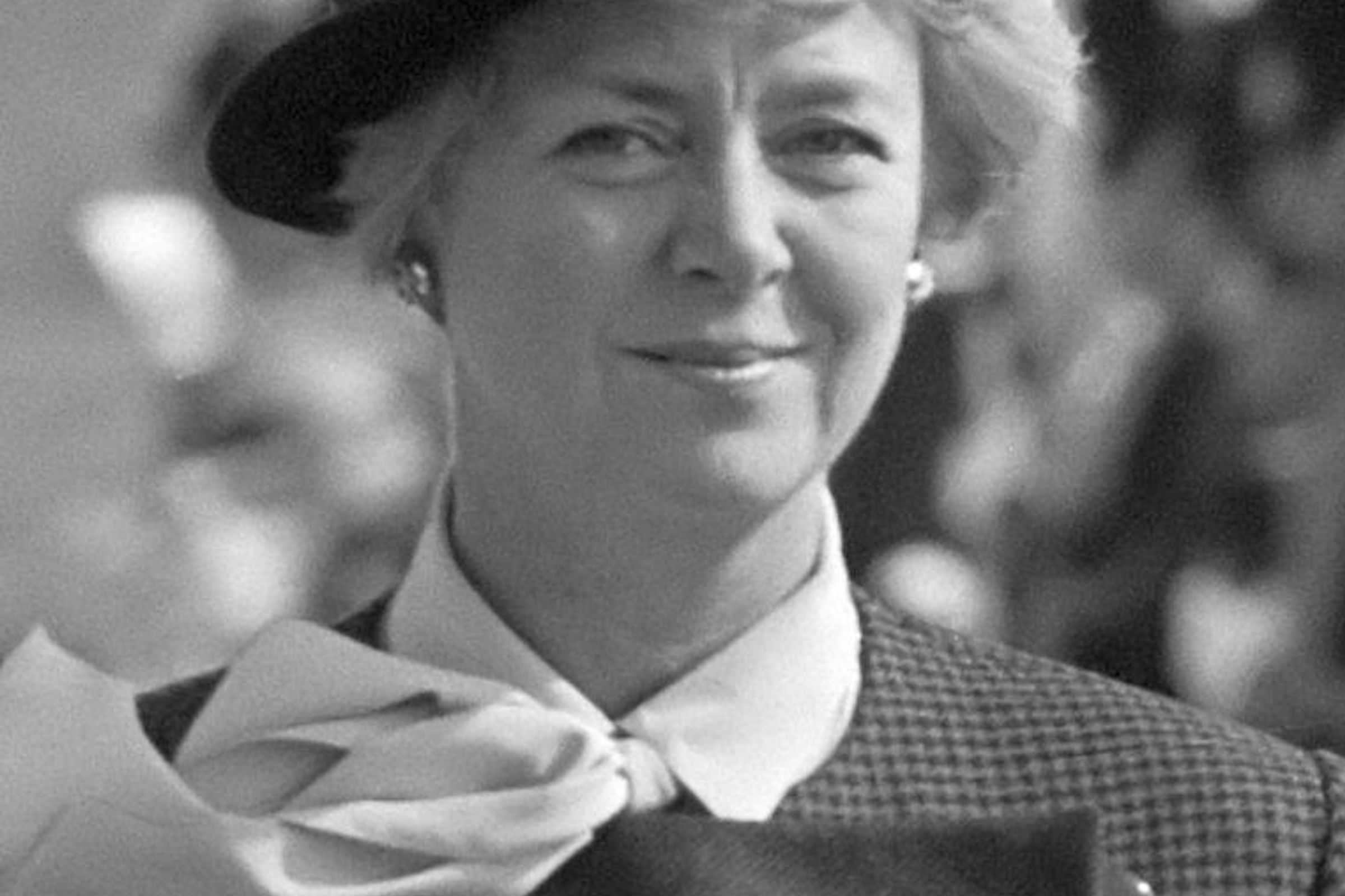 A black and white portrait of a smiling woman in a dark hat, white bow blouse, and patterned jacket.