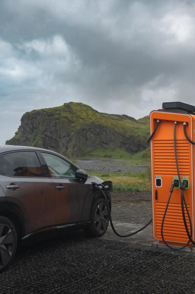 Electric car charging at an orange station in a rugged, mossy landscape.