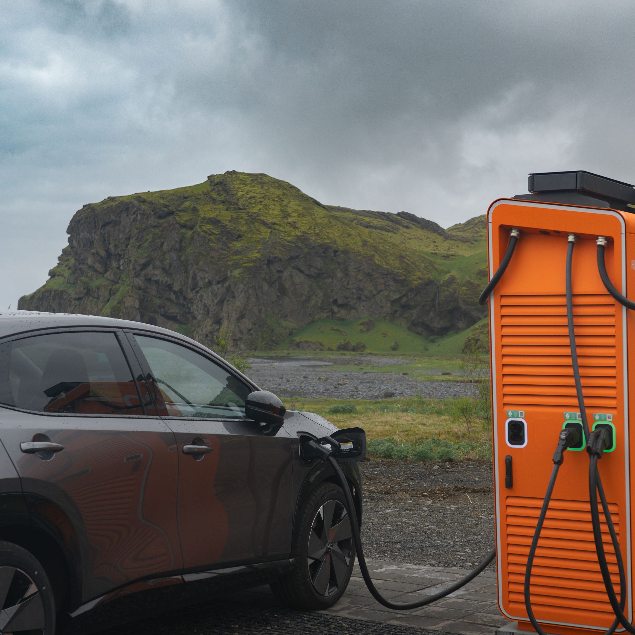 Electric car charging at an orange station in a rugged, mossy landscape.