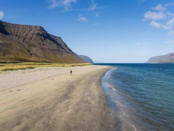 A person walks on a wide sandy beach beside a fjord, with large mountains under a clear blue sky.