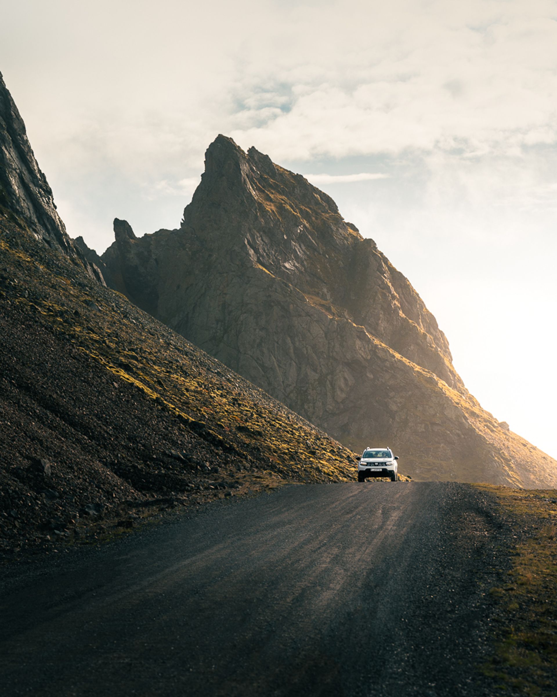 A white SUV on a gravel road winding through rugged, sunlit mountains.