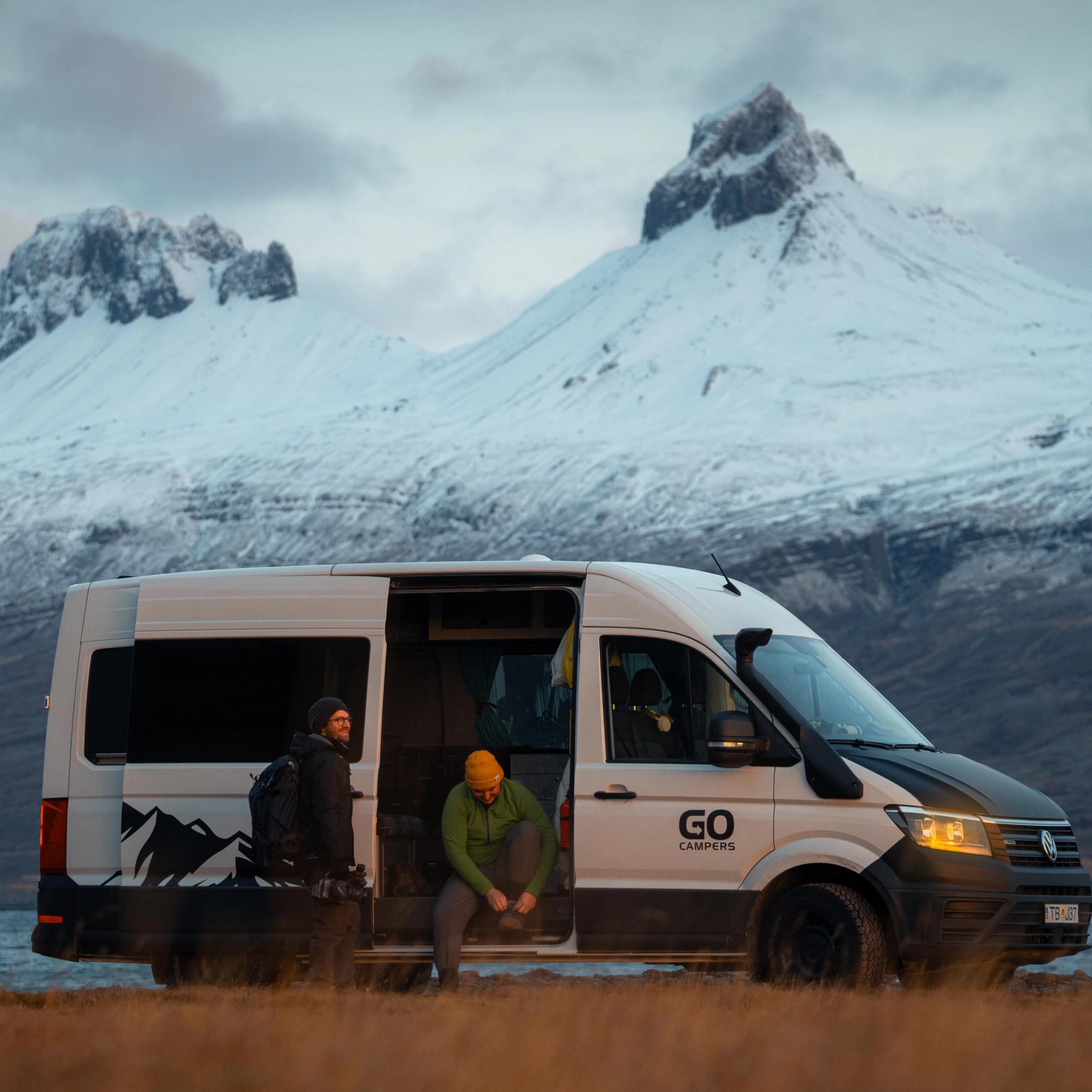 Two men are by a white and black camper van on a grassy field, with snow-capped mountains and a lake in the background.