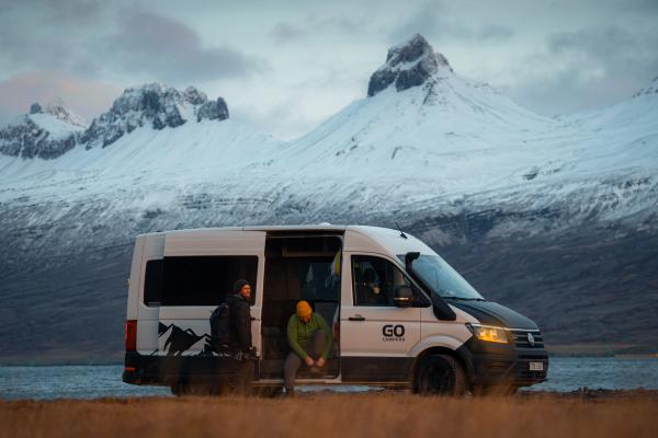 Dos personas junto a una furgoneta blanca de GO CAMPERS con la puerta lateral abierta; una de ellas se está atando un zapato, con un lago y montañas nevadas al fondo.