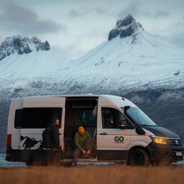 Una furgoneta camper blanca con dos personas junto a su puerta lateral abierta, estacionada junto a un lago con montañas nevadas en el fondo.