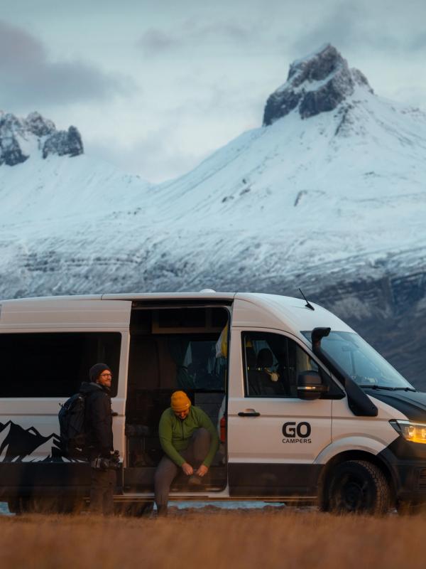 Two people with an open white camper van, one tying shoes, against snowy mountains and water.