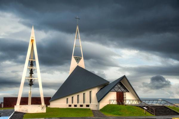 A modern, angular white church with a tall spire and a separate bell tower stands under dark, dramatic clouds.