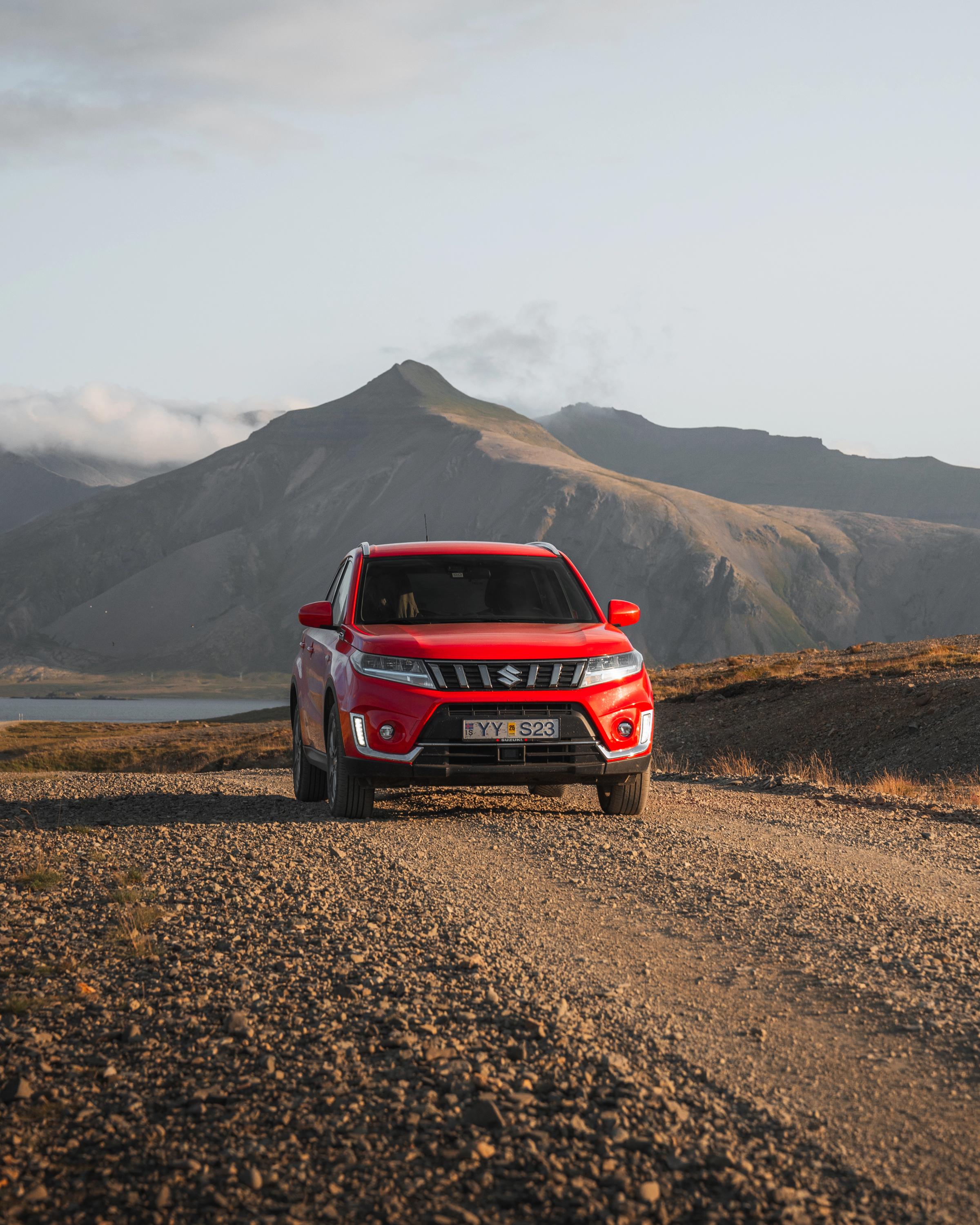 Vue de face d’un SUV Suzuki Vitara sur une route de gravier au bord de l’eau, avec des montagnes en arrière-plan