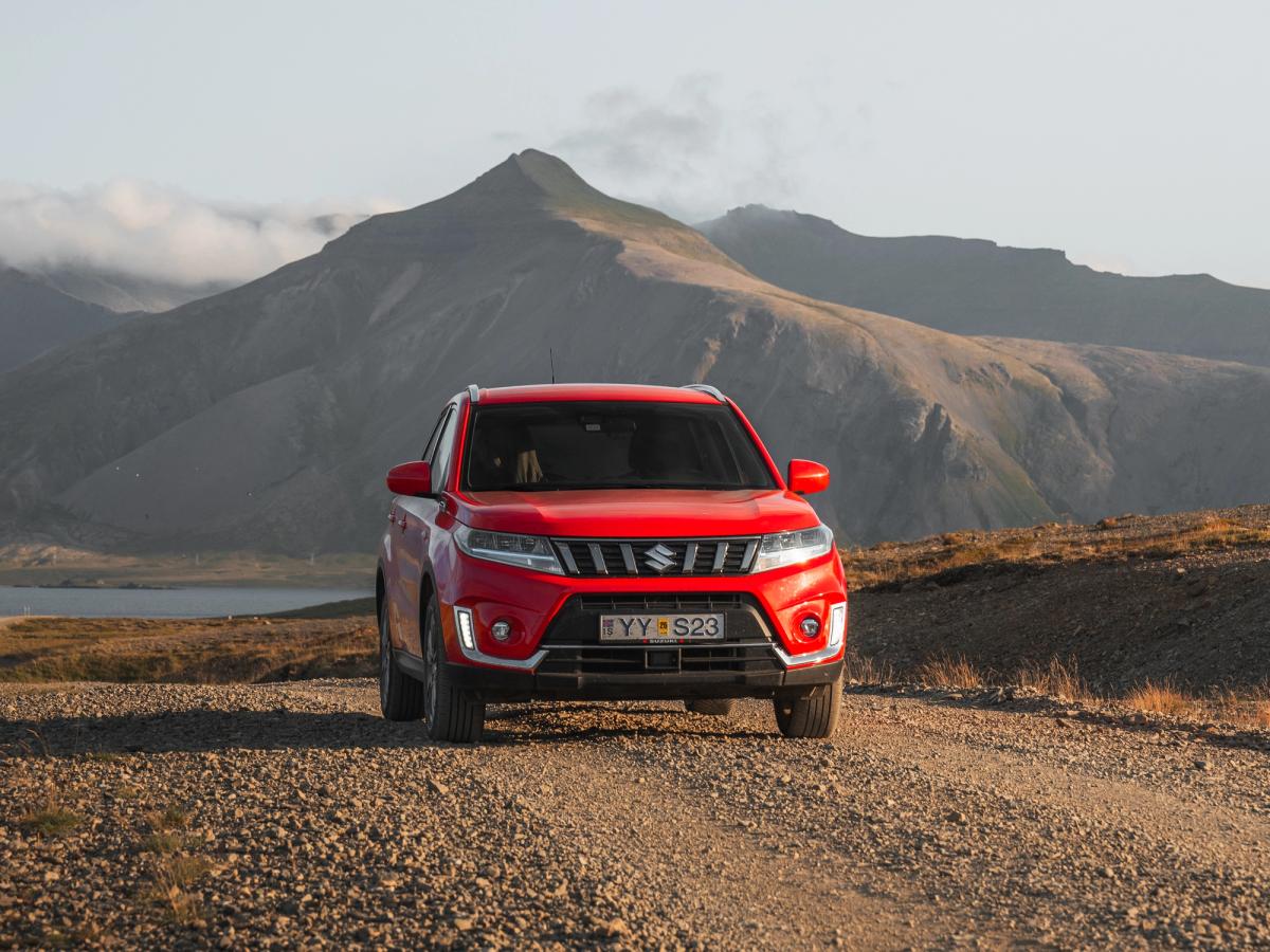 Vue avant du Vitara sur une route côtière Vue de face d’un SUV Suzuki Vitara sur une route de gravier au bord de l’eau, avec des montagnes en arrière-plan