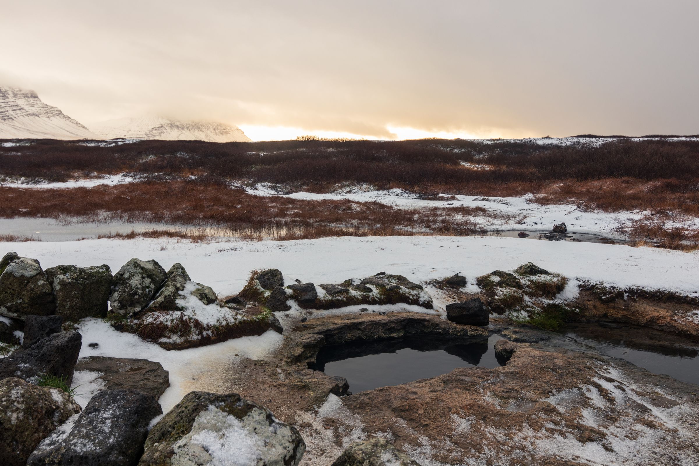 A natural hot spring surrounded by snow-dusted rocks in a winter landscape with distant mountains and a bright sky.
