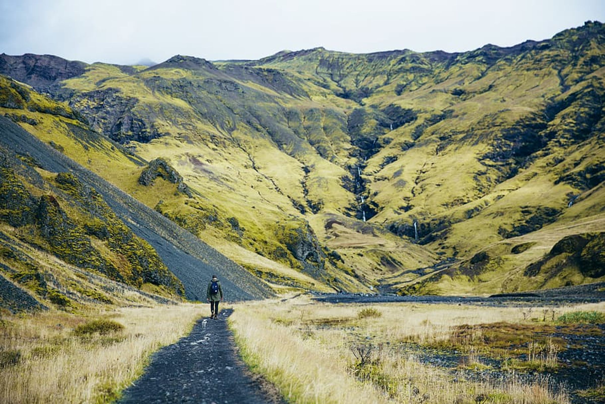 A person walks on a dirt path through a valley with green mountains and a waterfall.