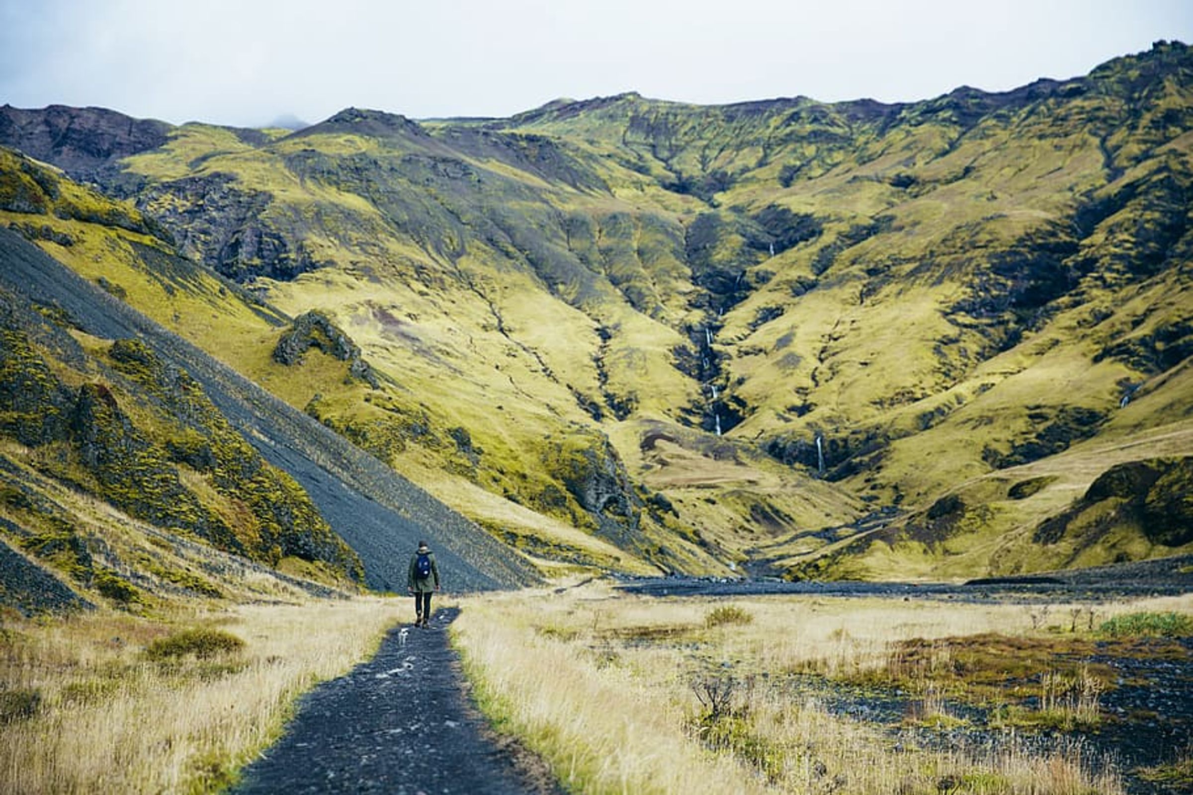A person walks on a dirt path through a valley with green mountains and a waterfall.