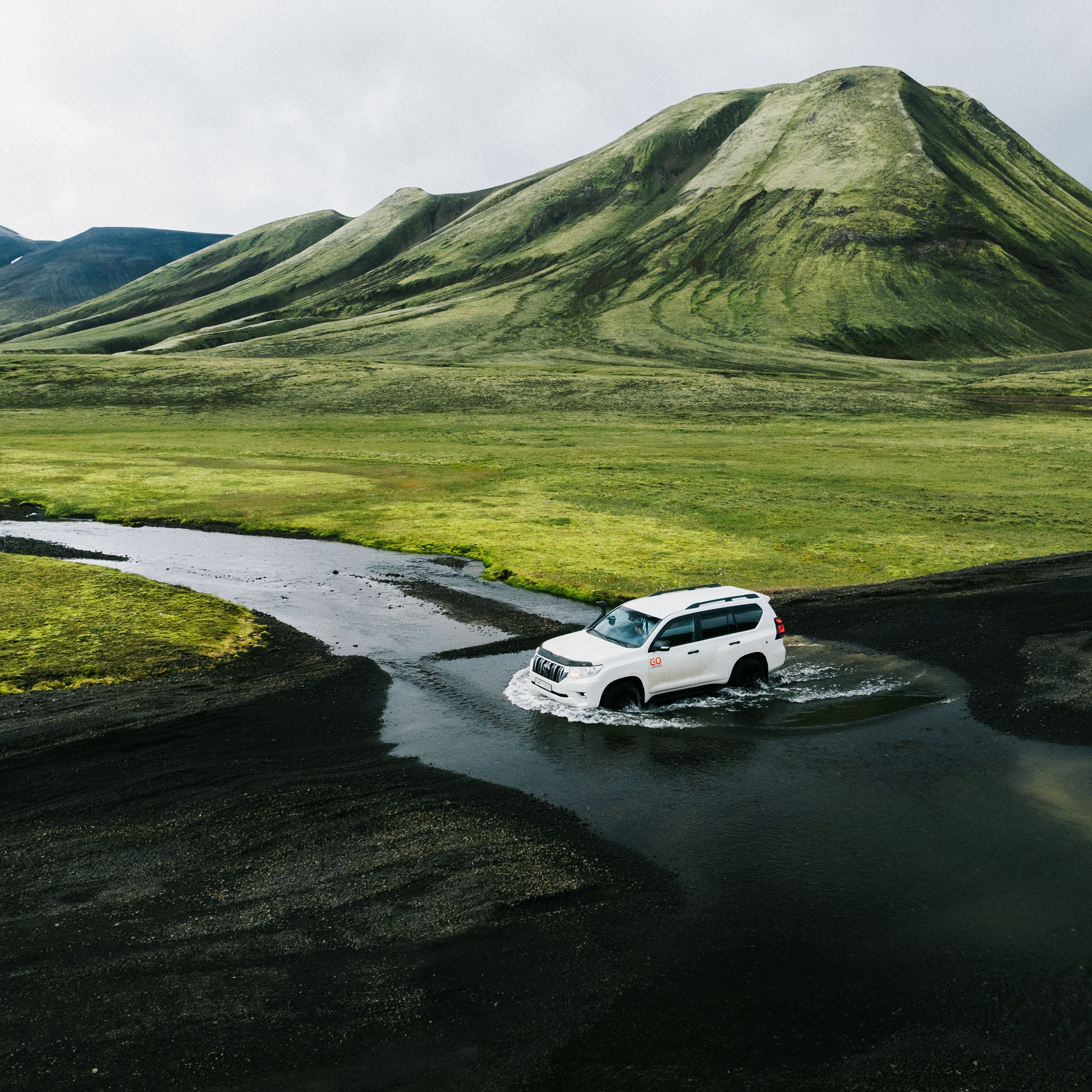 White SUV driving through a dark river in a landscape of green hills and black volcanic ground.