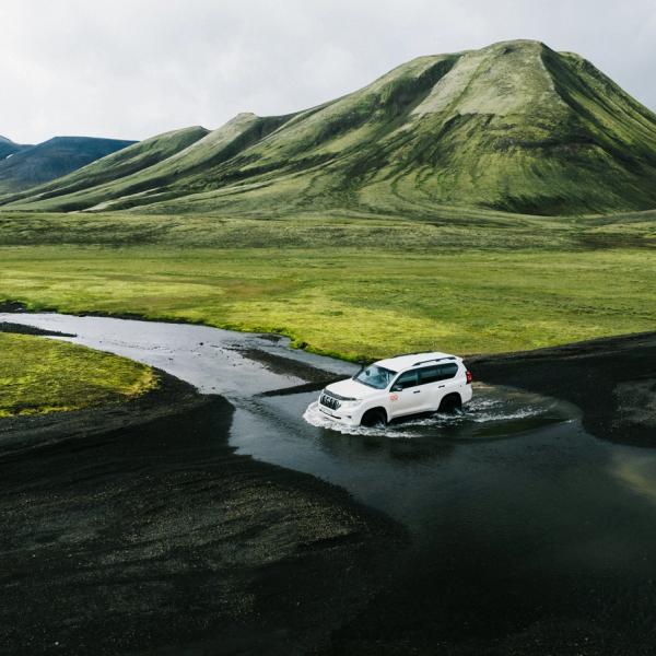 White SUV driving through a shallow river on dark volcanic terrain, with green mountains in the background.