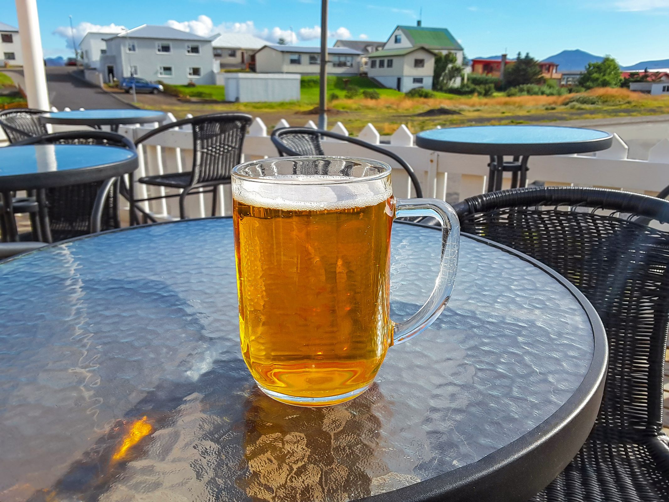 A mug of beer on a glass table at an outdoor cafe with houses in the background.