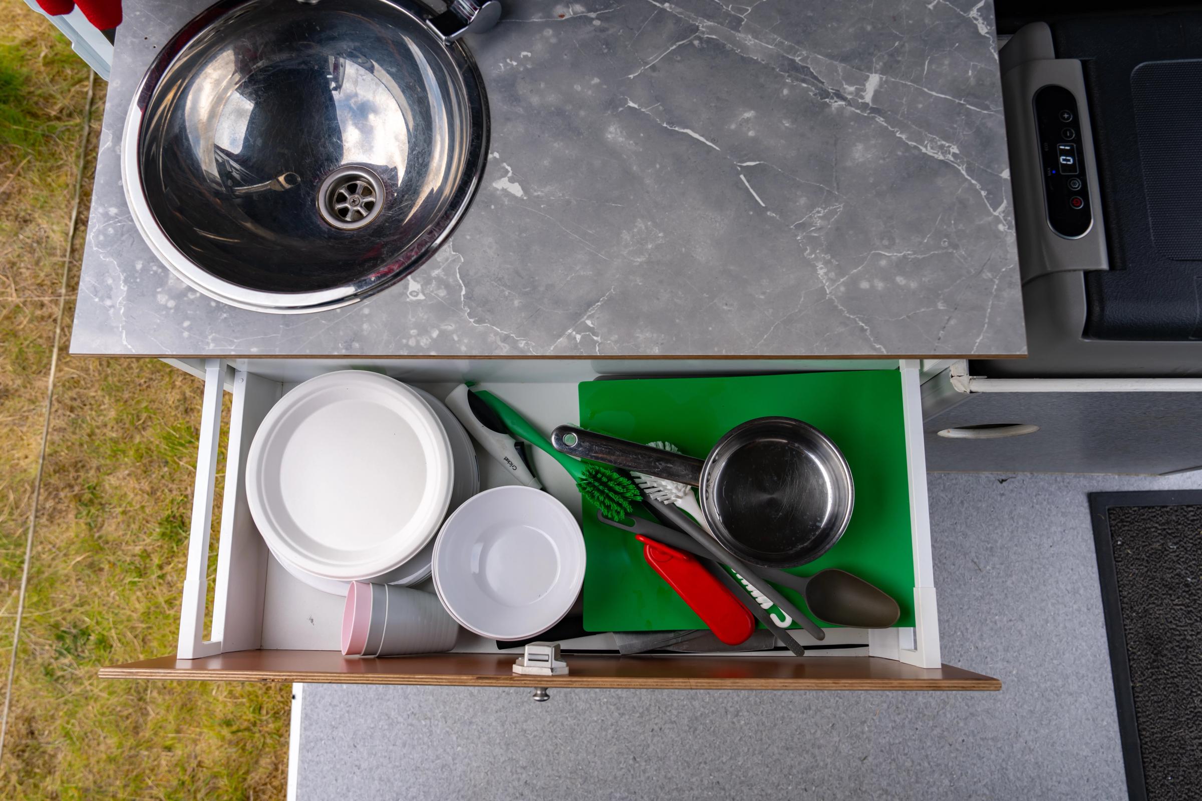Open kitchen drawer with plates, bowls, a pot and utensils below a steel sink