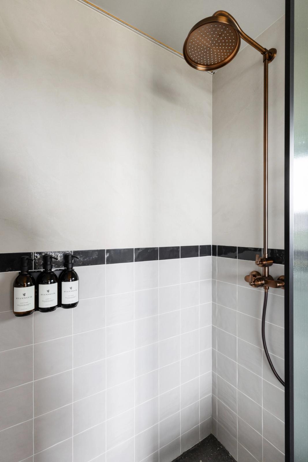 Modern shower with antique bronze fixtures, light grey tiles, a black border, and three wall-mounted soap dispensers.