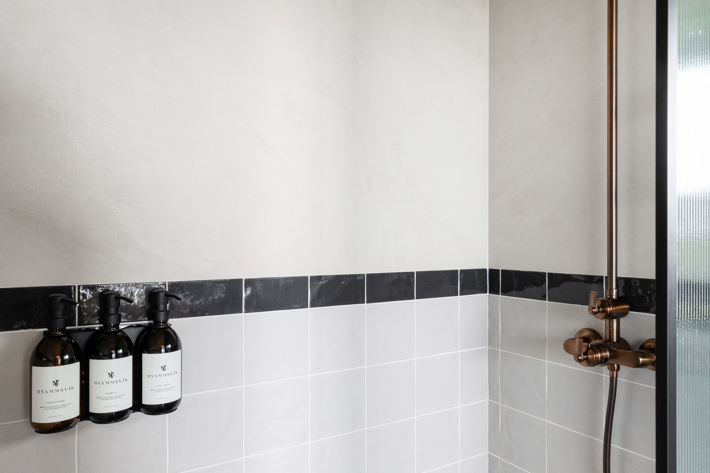 Modern shower with antique bronze fixtures, light grey tiles, a black border, and three wall-mounted soap dispensers.