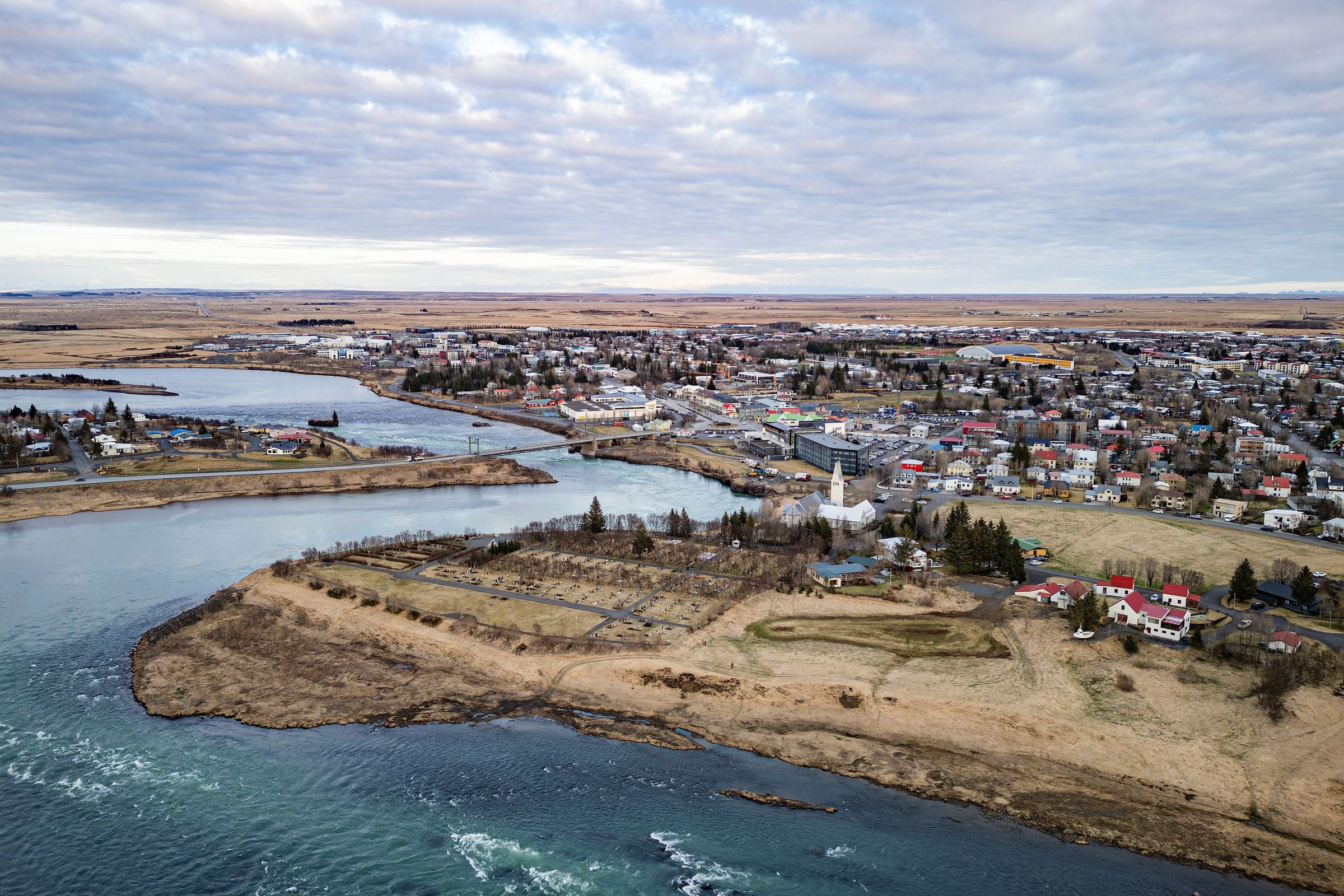 Aerial view of a town on a winding river with a bridge and a prominent white church spire, surrounded by dry fields under a cloudy sky.