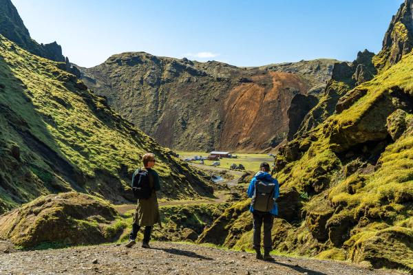 Two hikers with backpacks overlook a green, mossy valley with a camp and colorful mountain in the distance.