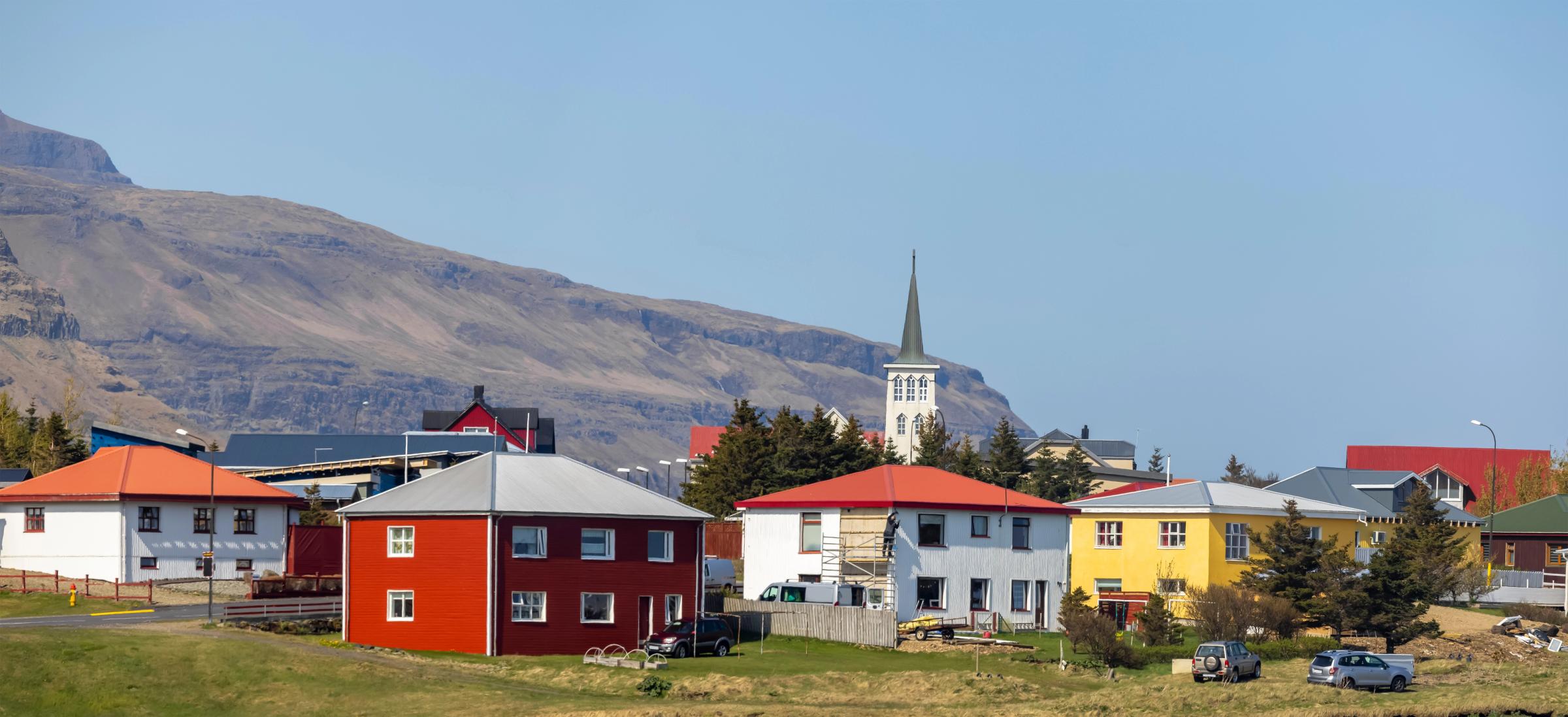 A small town with colorful houses and a white church steeple against a large mountain range under a clear blue sky.