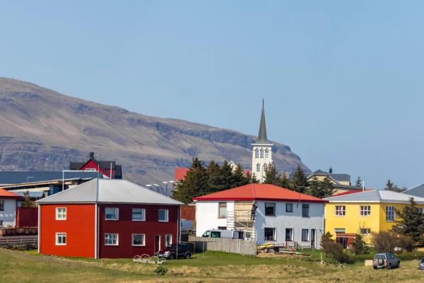 A small town with colorful houses and a white church steeple against a large mountain range under a clear blue sky.