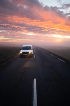 A white van drives on a foggy road under a vibrant orange and pink sunset sky.