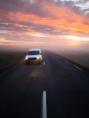A white van drives on a foggy road under a vibrant orange and pink sunset sky.