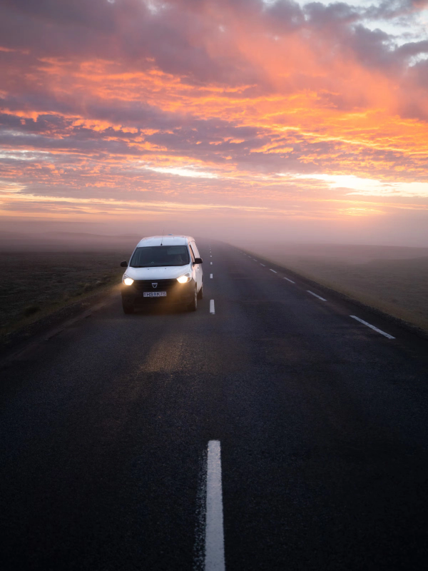 A white van drives on a foggy road under a vibrant orange and pink sunset sky.