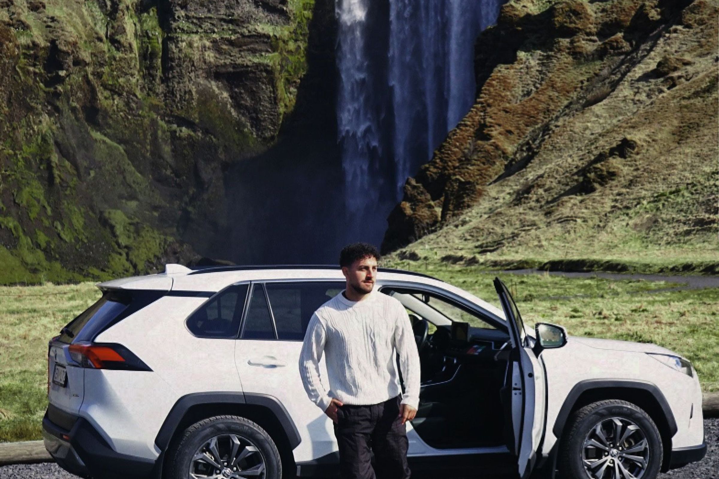 a man standing next to a white suv in front of a waterfall