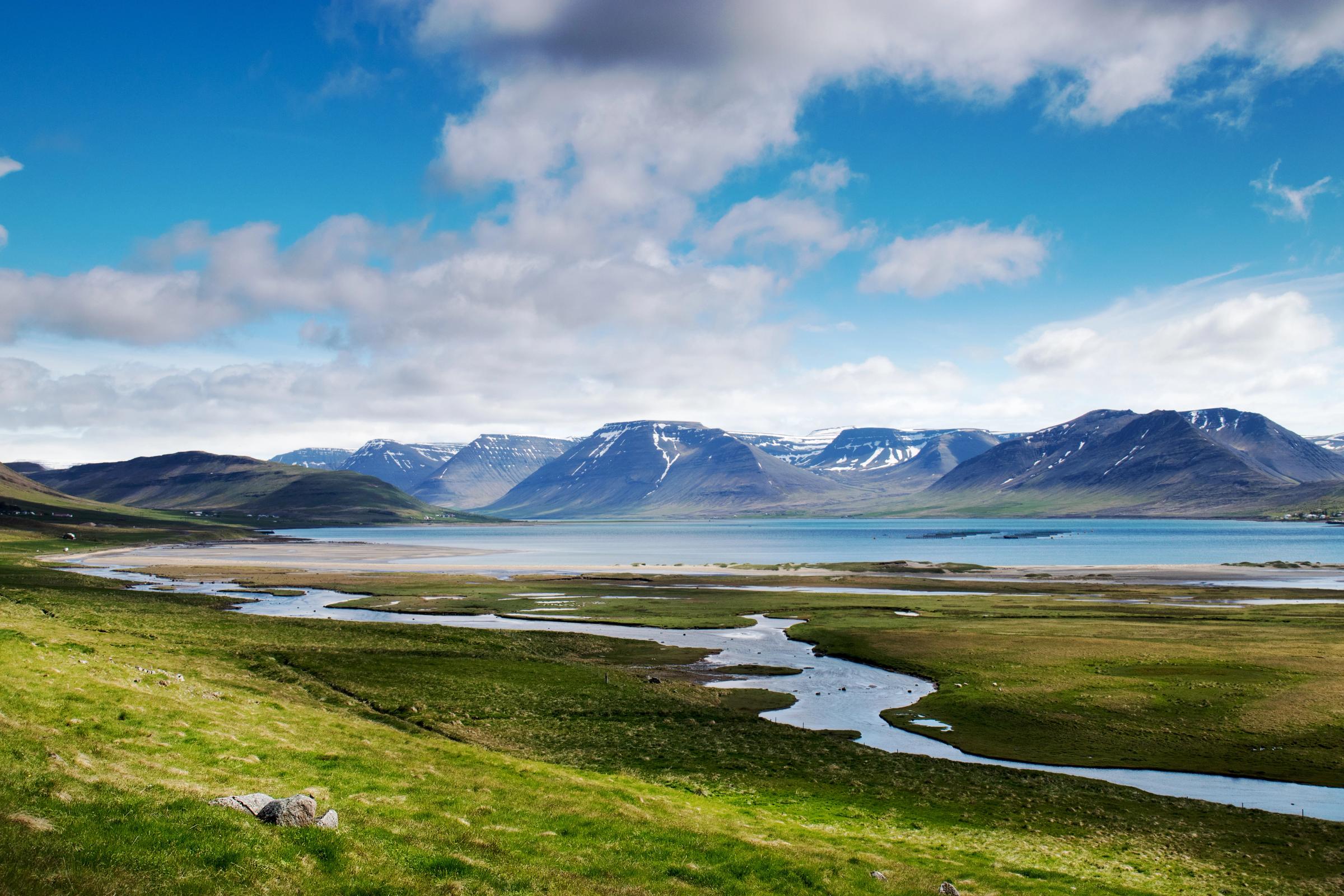Un río serpenteante atraviesa campos verdes y desemboca en un fiordo, rodeado de montañas nevadas bajo un cielo azul con nubes.