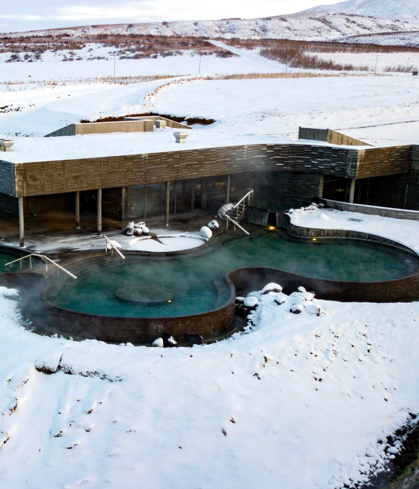 Aerial view of steaming outdoor geothermal pools surrounded by snow, with a modern building and distant hills.