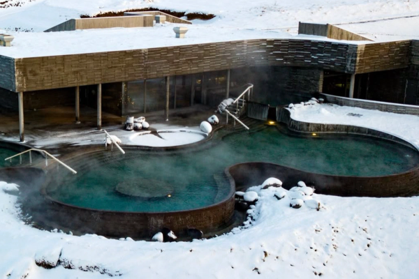 Piscinas geotérmicas humeantes al aire libre, rodeadas de nieve, junto a un edificio moderno y colinas a lo lejos.