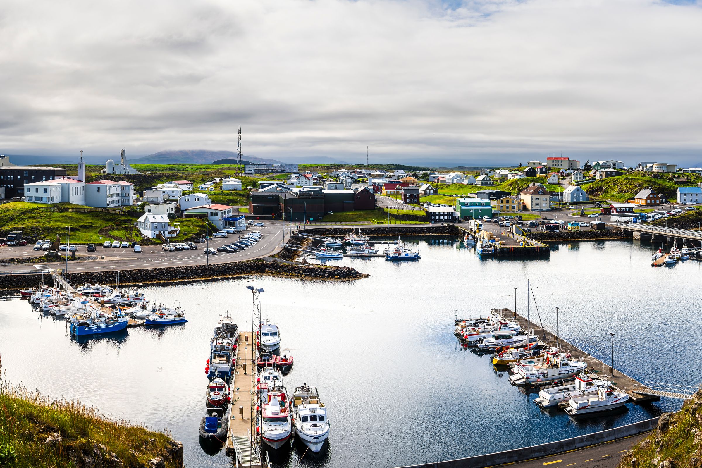 Panoramic view of a coastal town with a busy harbor filled with boats, surrounded by green hills under a cloudy sky.