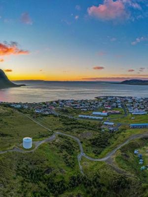 Panoramic aerial view of a town on a bay, framed by green mountains, with a prominent peak and a colorful sunset sky.