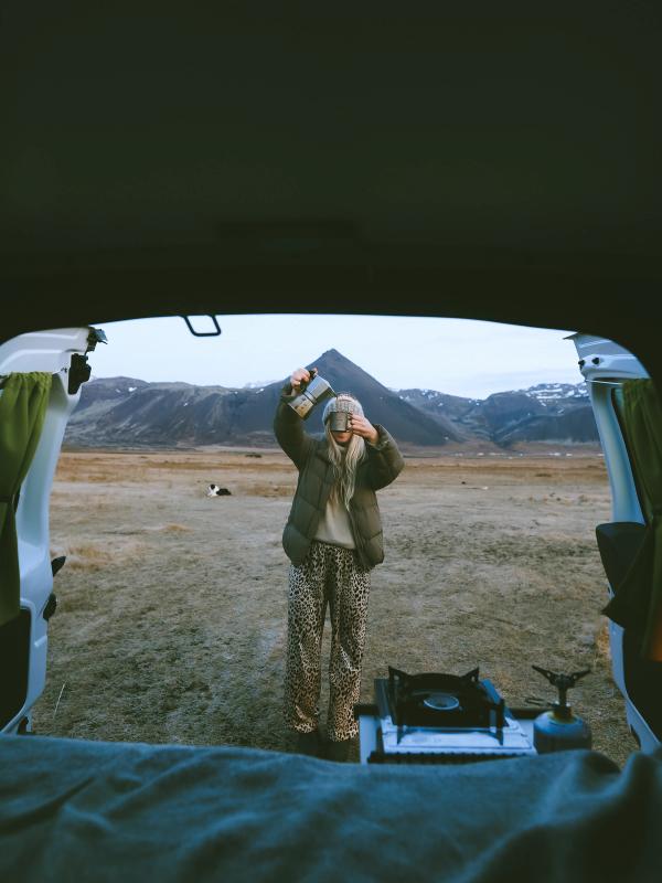 View from inside a camper van of a person pouring coffee outdoors in a rugged mountain landscape with a camp stove.