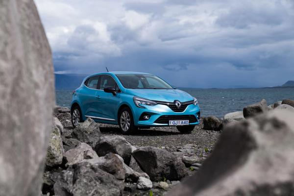 A light blue Renault Clio parked on a rocky shore by water under a cloudy sky.