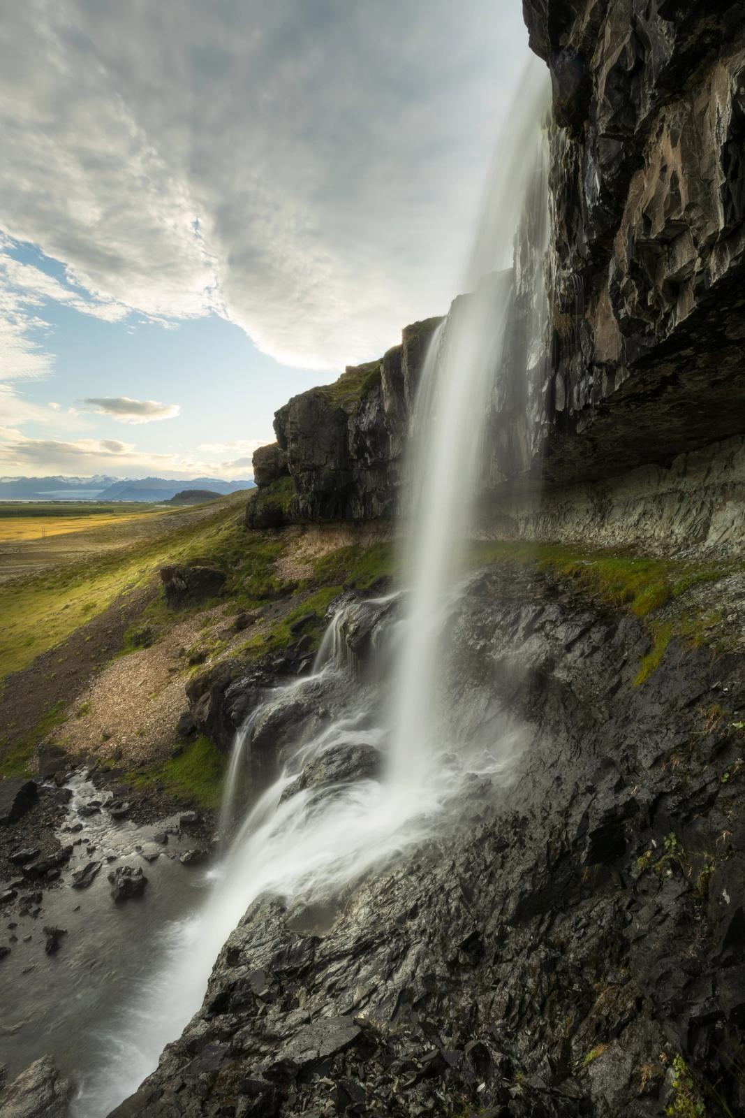 A long exposure of a waterfall cascading down a dark cliff, with a green landscape and cloudy sky.
