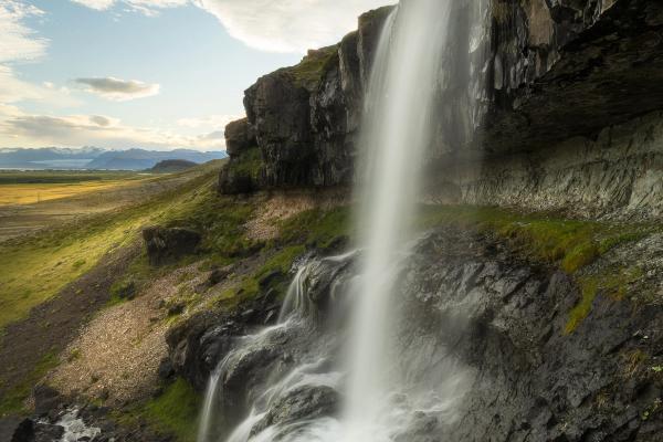 A long exposure of a waterfall cascading down a dark cliff, with a green landscape and cloudy sky.