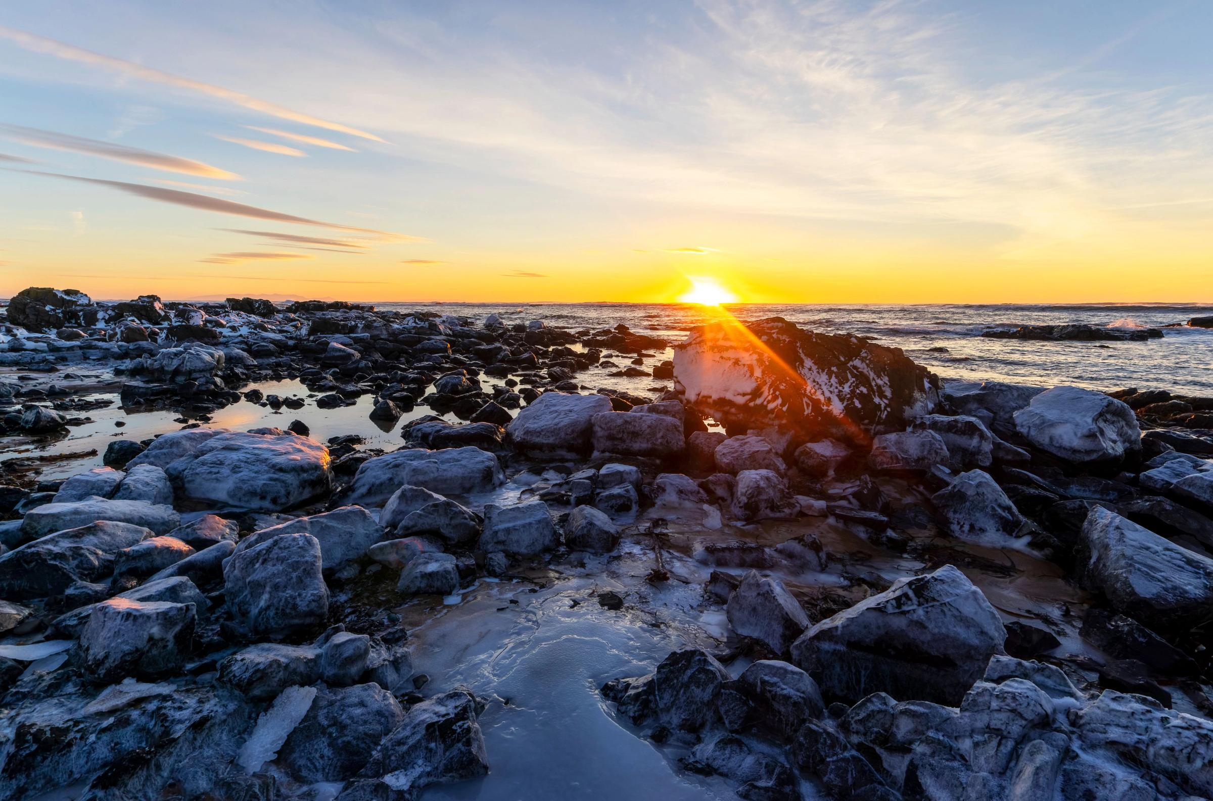 the sun is setting over the ocean behind a rocky shoreline .