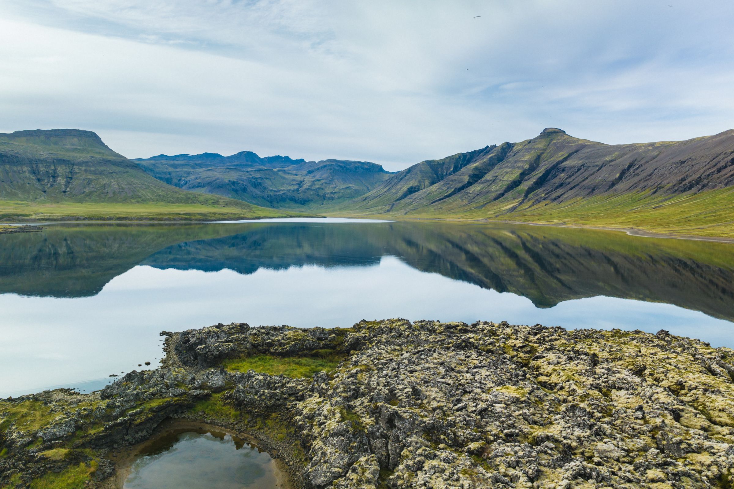 A calm lake reflects green mountains under a cloudy sky, with rocky, mossy ground in the foreground.