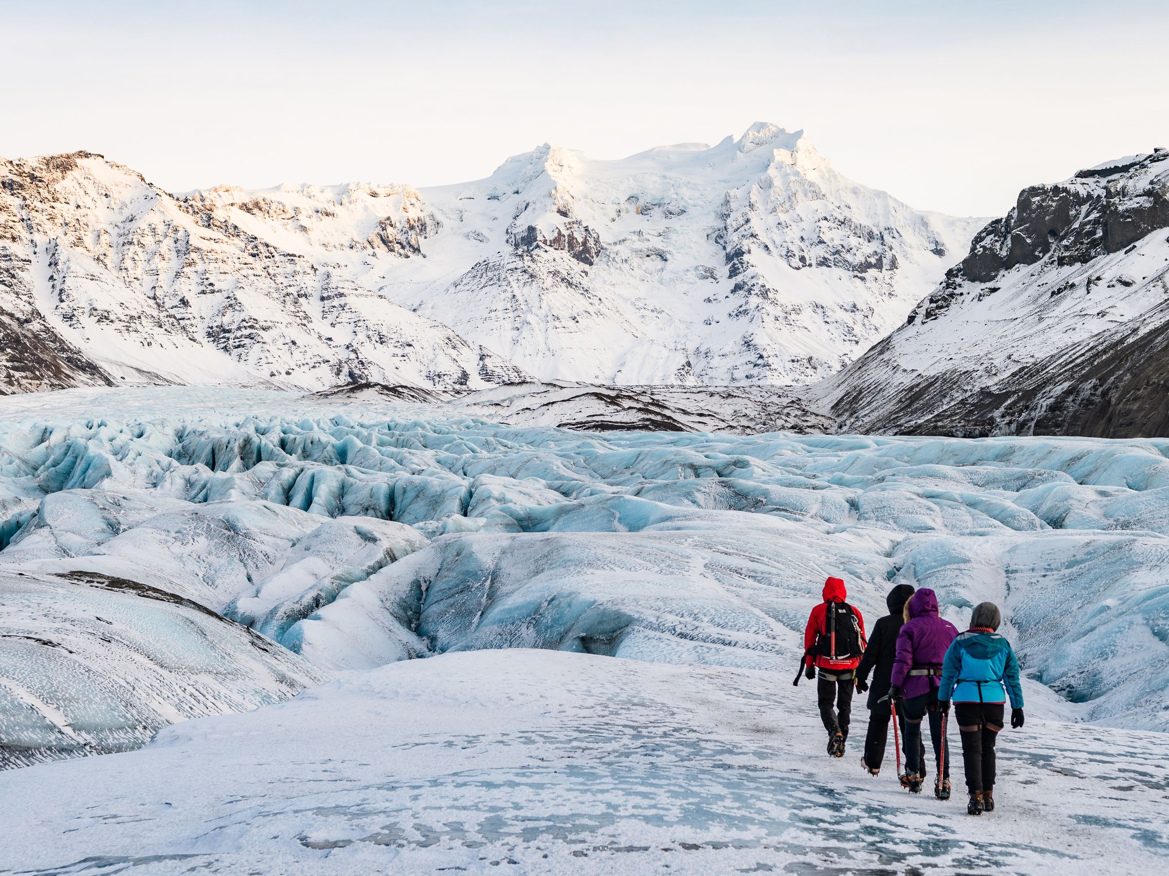 Four people hike across a blue glacier with snow-capped mountains in the background.