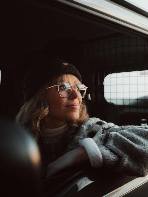 A woman in a beanie and glasses looks out a car window.